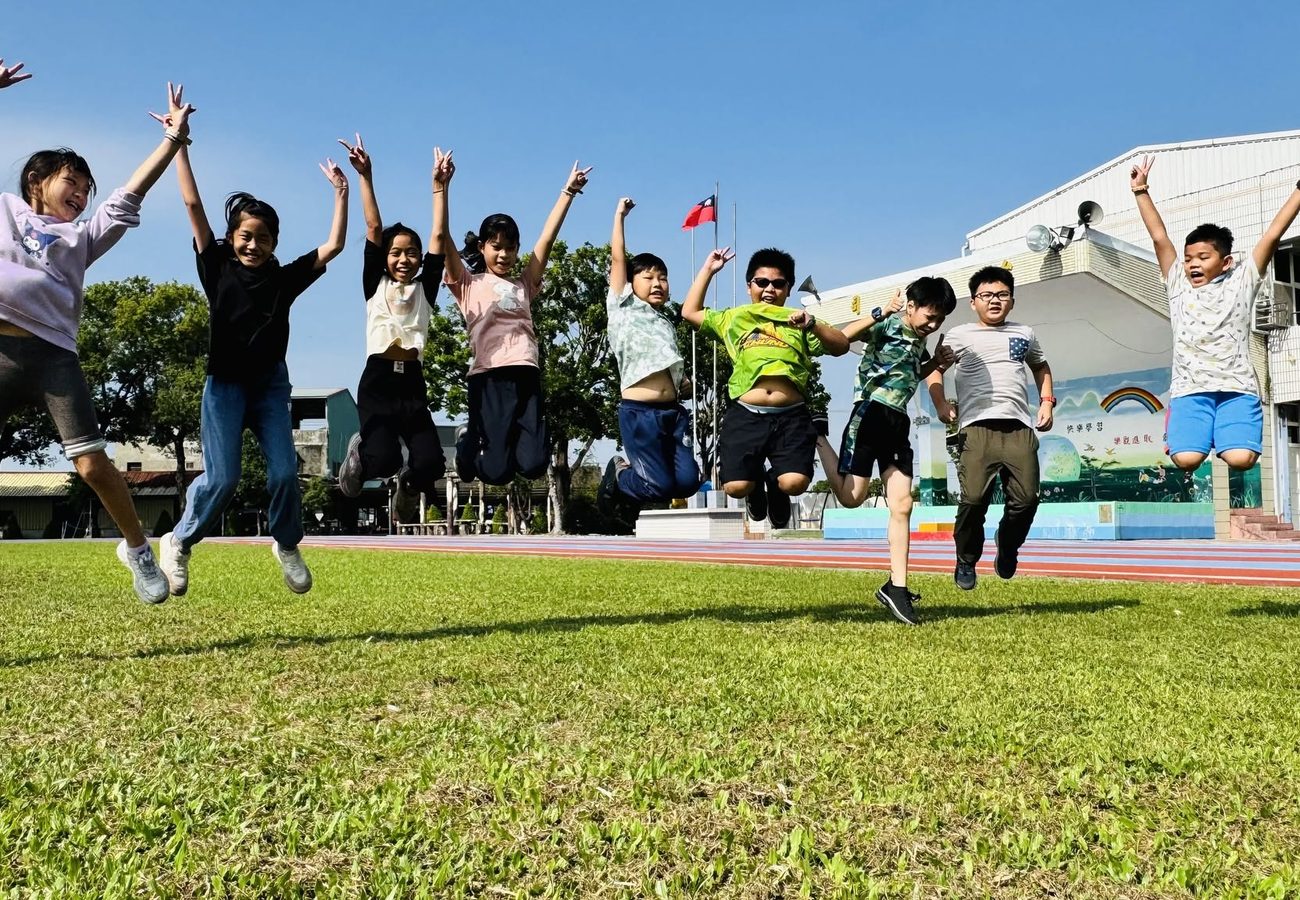 朝興國小學生在草地上開心跳躍、比出勝利手勢，背後是司令台的彩虹願景牆 Chaohsing children leaping on the grass with peace signs, the rainbow vision mural on the stage wall behind them