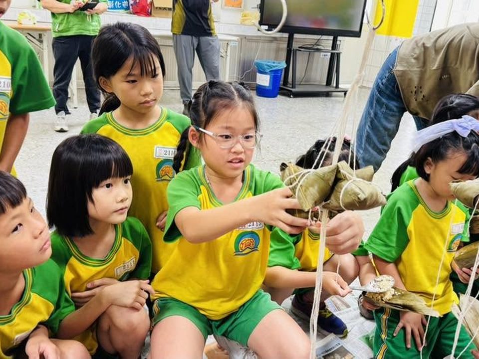 朝興國小學生圍在一起，用棉繩綁製竹葉粽子，動手體驗傳統食物 Chaohsing children gathered around, tying bamboo-leaf rice dumplings with string in a hands-on food activity