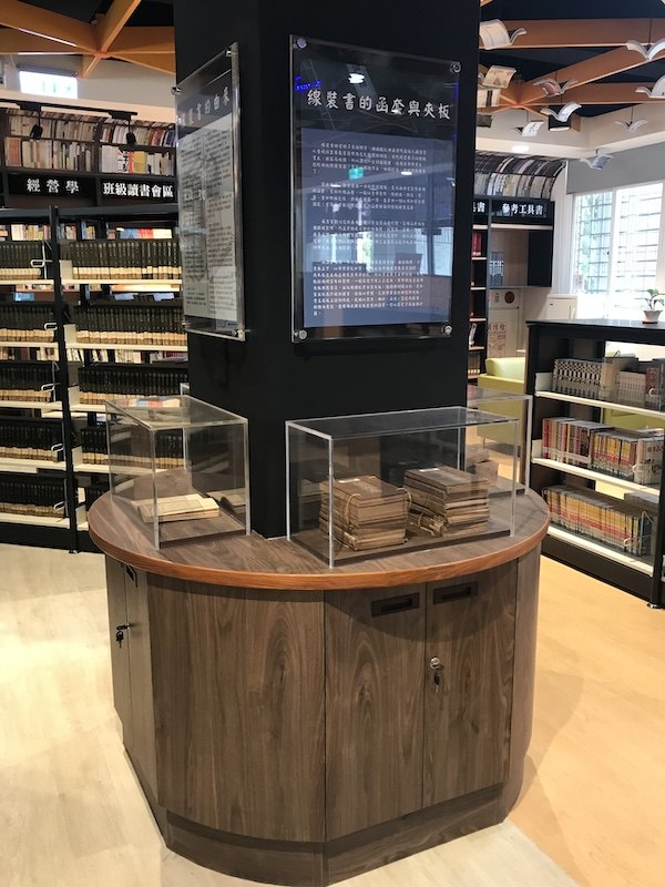 黑色柱體四面裝著壓克力海報架，底下是圓形木質展示桌與玻璃展櫃 A black pillar with acrylic poster frames on every face, standing on a round wooden display table with a glass case