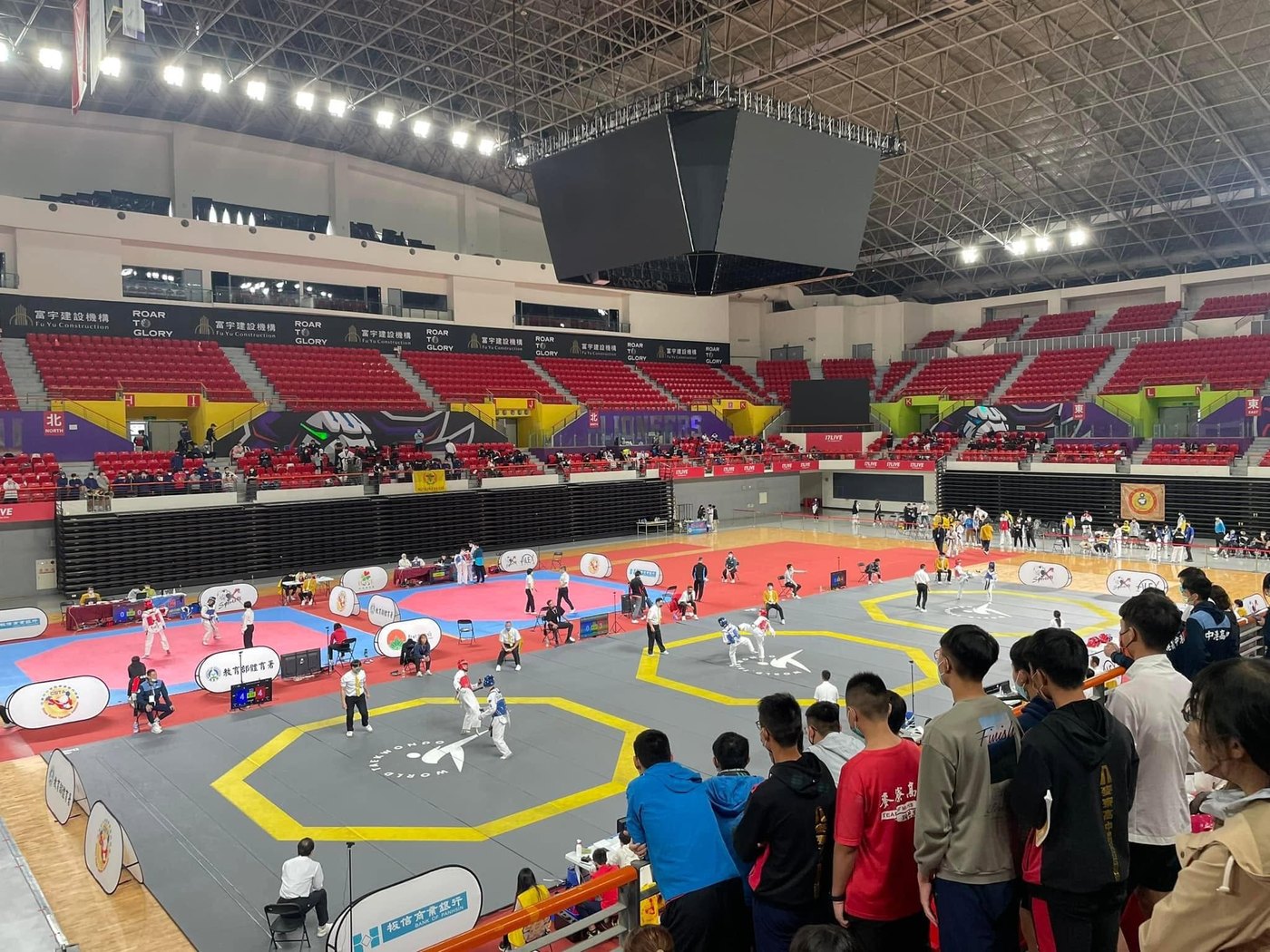 錦標賽場館全景，館內同時進行多場比賽，看台圍繞四周 A wide view of the championship venue with several contests running at once and stands all around