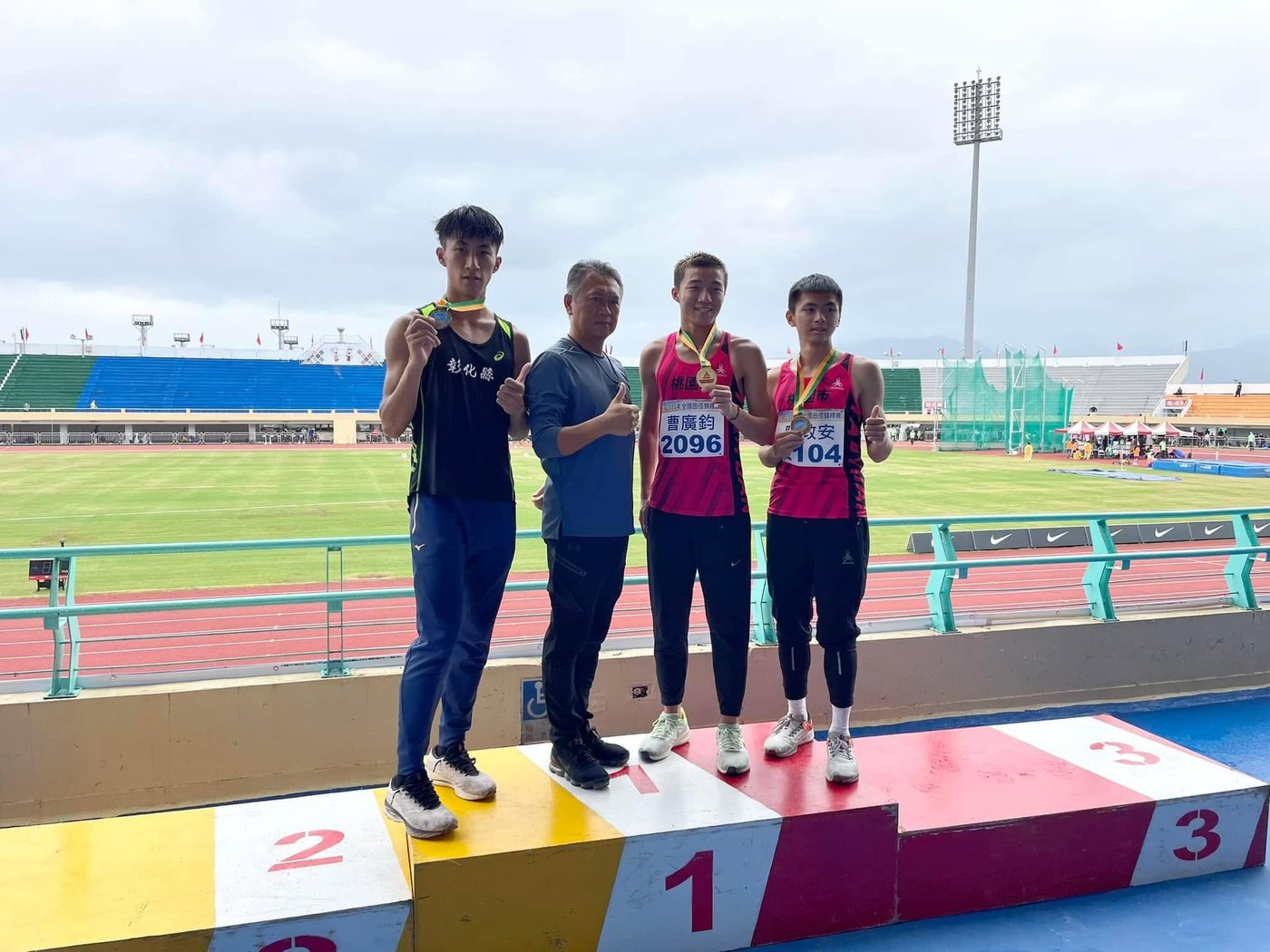 田徑隊員與師長在比賽頒獎台上合影，背後是跑道與看台 Track team members and staff on the medal podium at a meet, the track and stands behind them