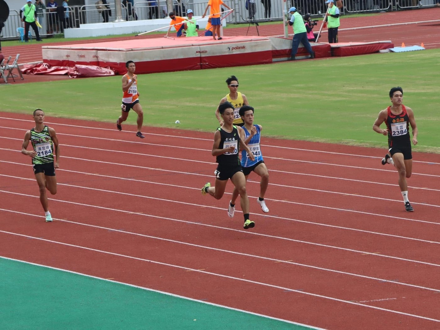 田徑隊員在紅色跑道上競速，場中是綠色草皮 Runners racing down the red track with the green infield beside them