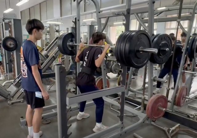 一名隊員在深蹲架前舉起槓鈴做深蹲，隊友在一旁觀察保護 A team member squatting under a loaded barbell in the weight room while a teammate spots