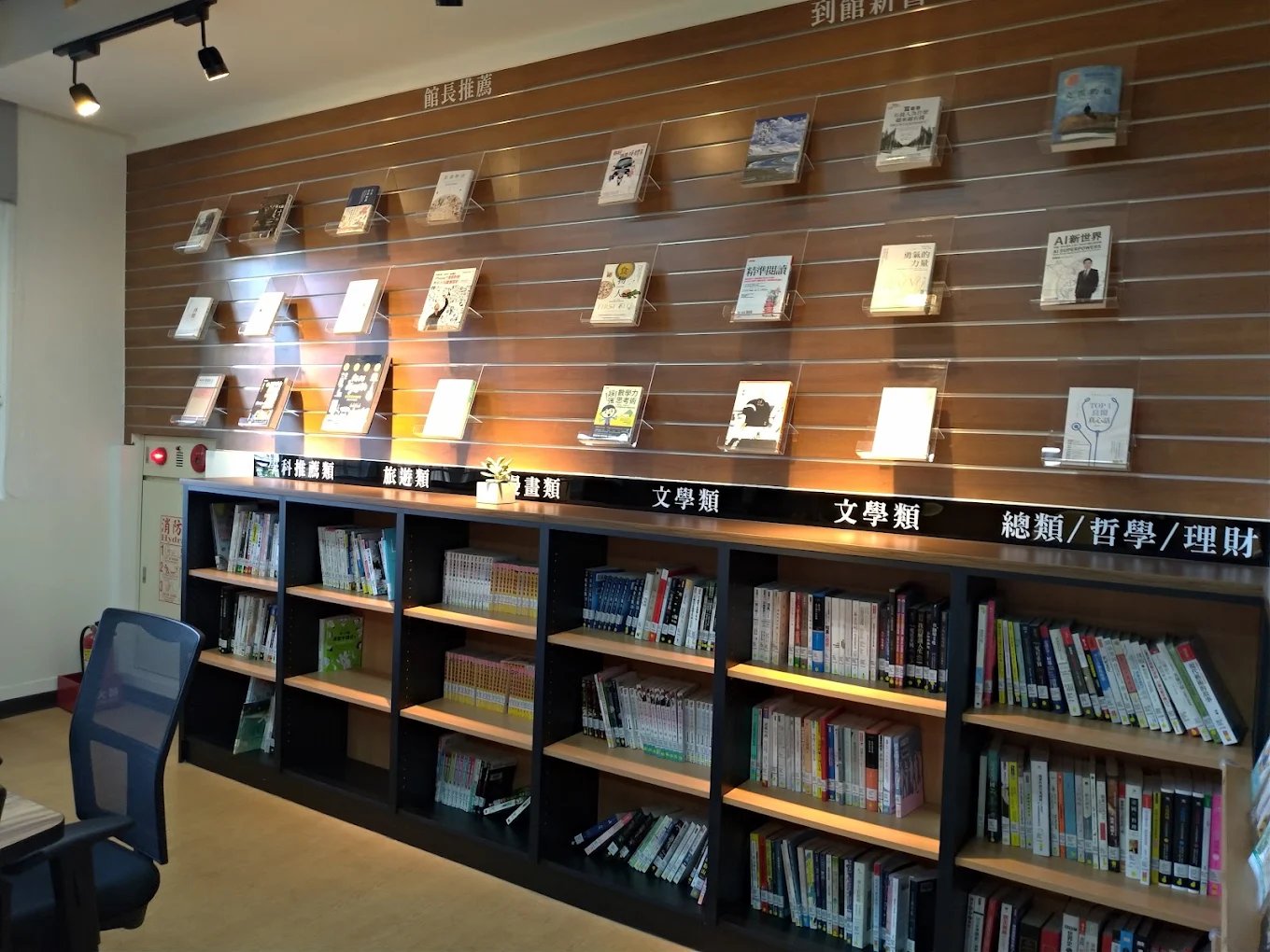 崇實圖書館的新書展示牆，木紋牆面上以壓克力書架展示「館長推薦」與「到館新書」 The new-arrivals wall at the ChungShih library, books displayed face-out on acrylic shelves under the headings for the librarian's picks and newly arrived titles
