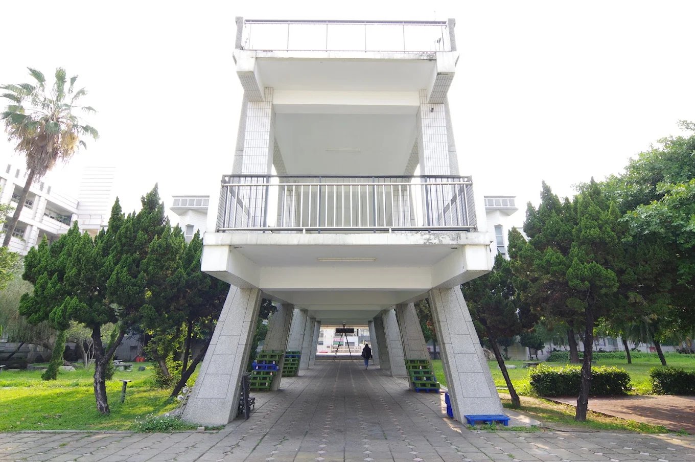 崇實高工校園中一座架高的穿堂建築，兩側種著整齊的柏樹，中央是磚砌步道 A raised portal building at ChungShih framed by clipped conifers, a paved walkway running straight through underneath
