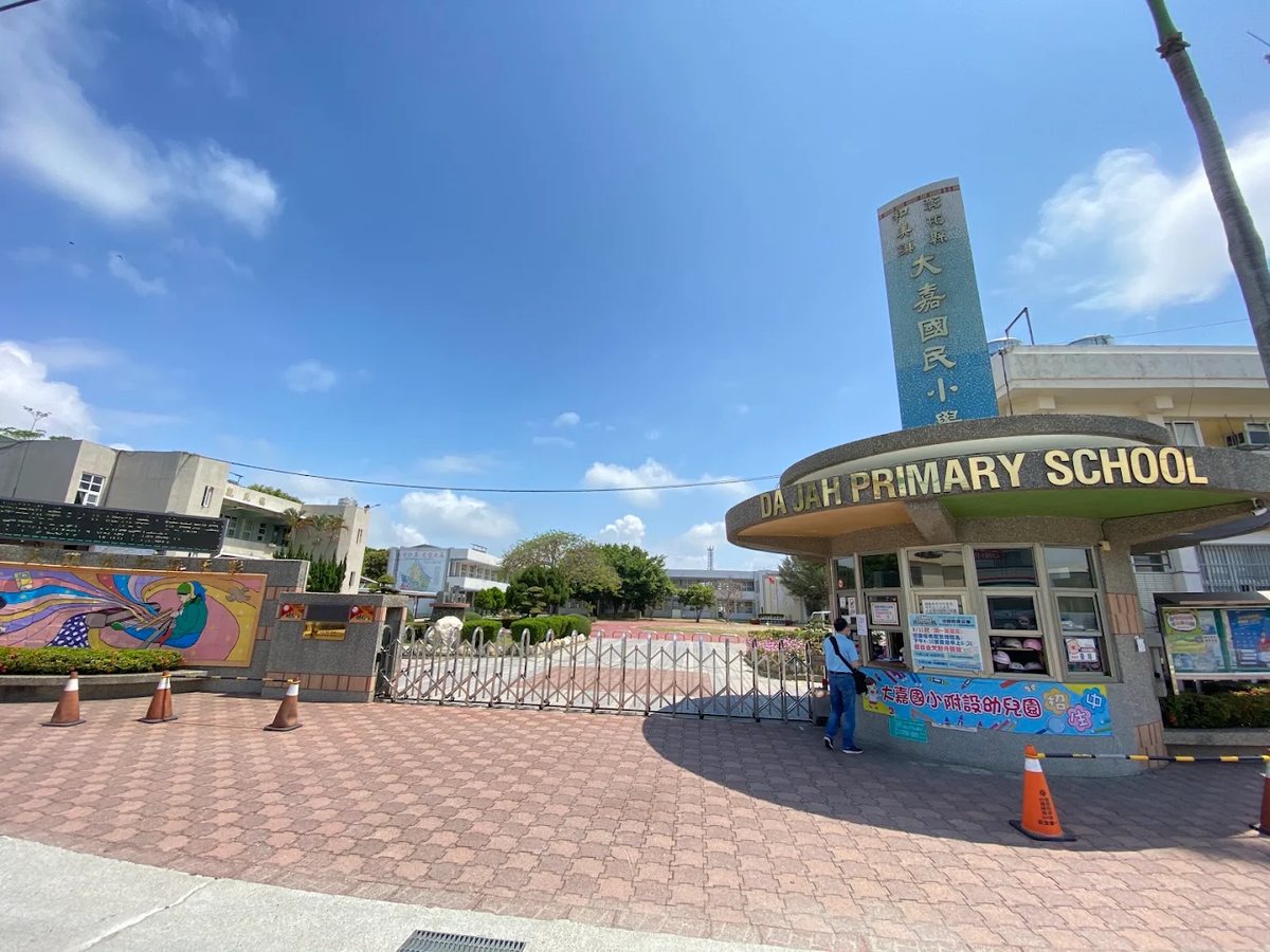 大嘉國小校門，金色英文校名 DA JAH PRIMARY SCHOOL 與中文校名塔柱，藍天白雲 The front gate of Da Jah Elementary, the school name in gold English letters beneath the Chinese name tower, under a blue sky