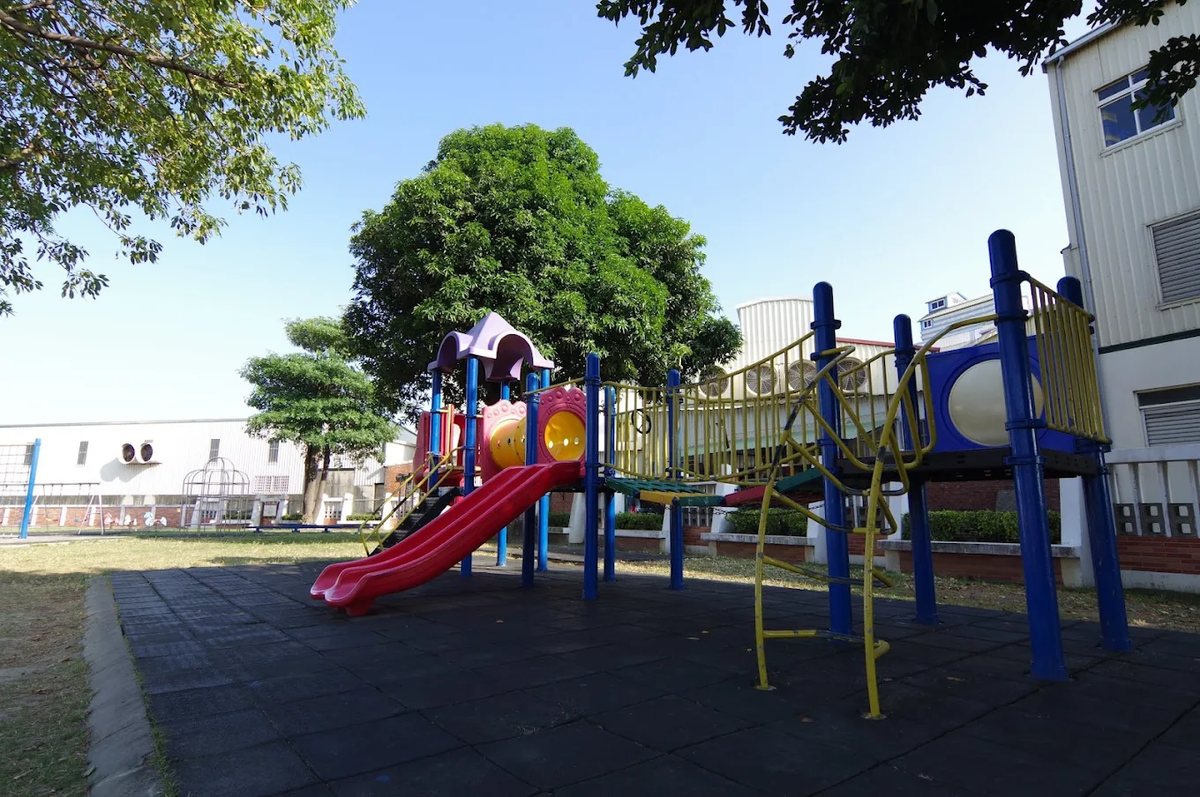 大嘉國小色彩鮮豔的遊戲場：紅色溜滑梯、藍色與黃色的攀爬架，大樹與藍天在後方 The brightly coloured playground at Da Jah — a red slide, blue and yellow climbing frames, a big tree and blue sky behind