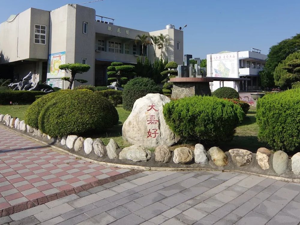 校園前庭一顆刻著紅色「大嘉好」三字的白色巨石，四周是修剪整齊的綠籬與校舍，藍天無雲 A white boulder in the front courtyard carved with the three red characters 大嘉好, trimmed hedges and school buildings around it under a clear blue sky