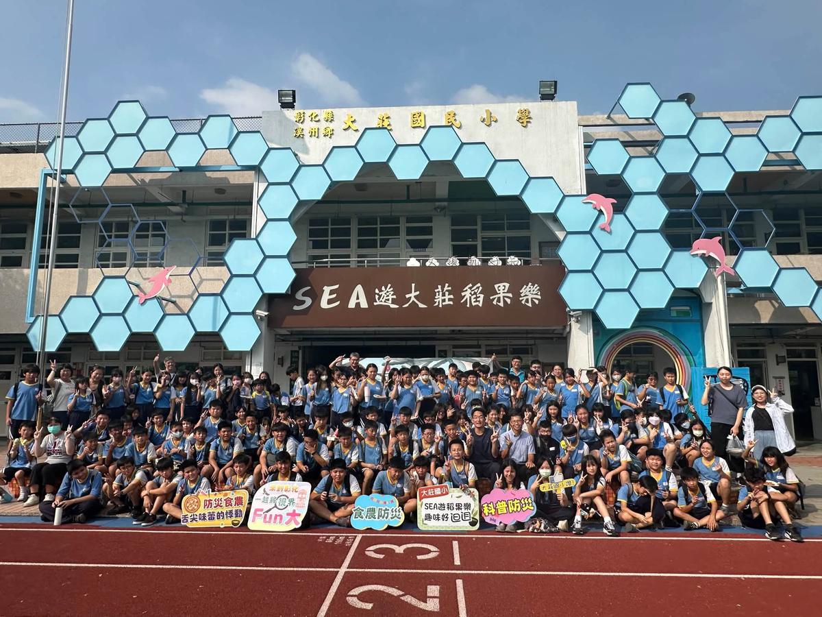 Students from Dingfan and Dajuang Elementary pose for a big group photo in front of Dajuang's hexagon-patterned school gate, holding signs about disaster prevention, food and farming, and science.