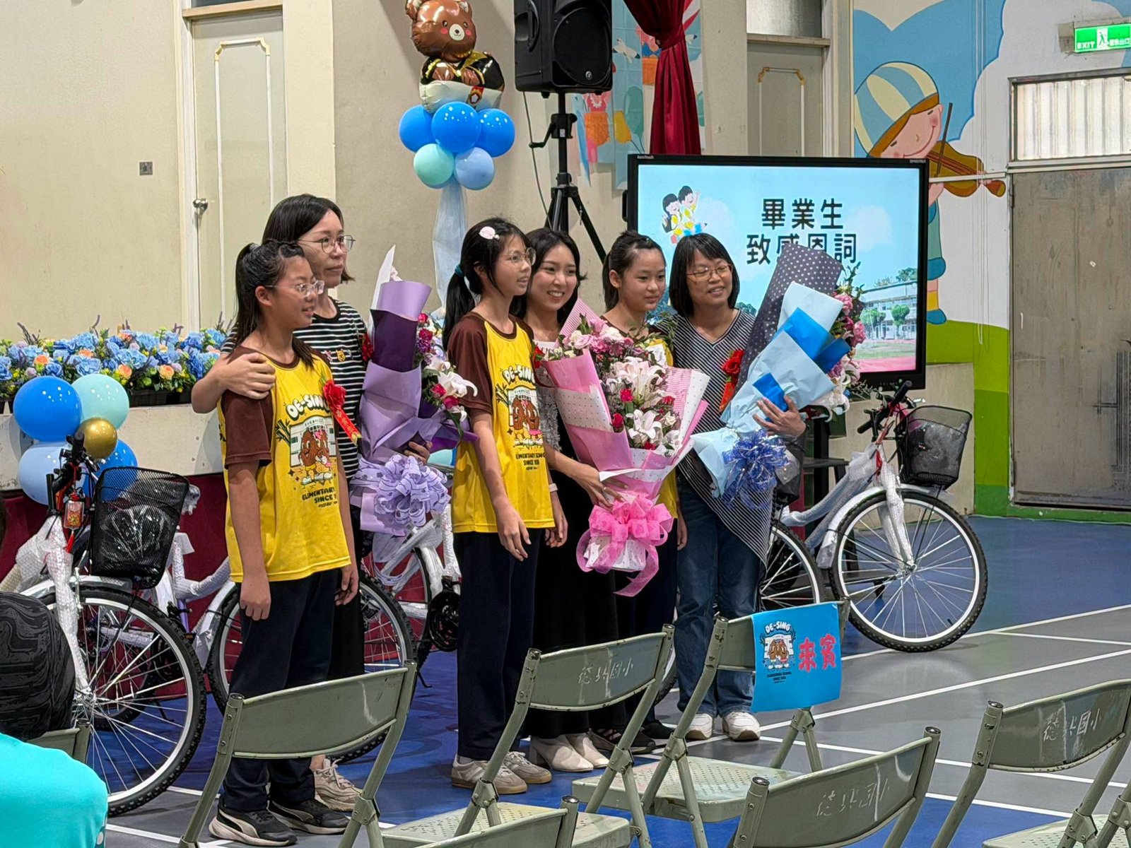 Graduates holding flower bouquets on stage beside gifted bicycles