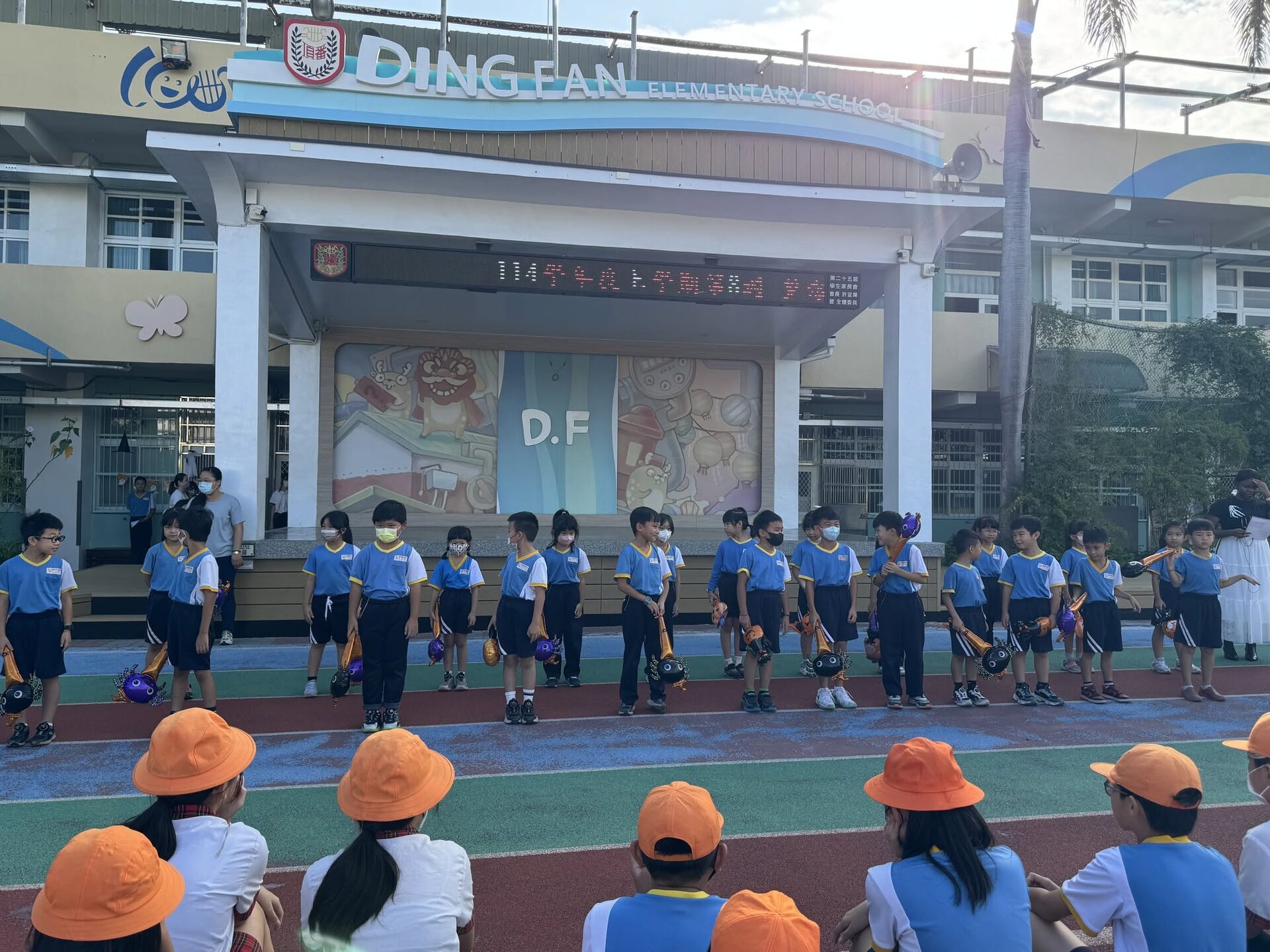 Dingfan students lined up in front of the school's Ding Fan Elementary School sign holding Halloween props 頂番學生在校門口拿著萬聖節道具