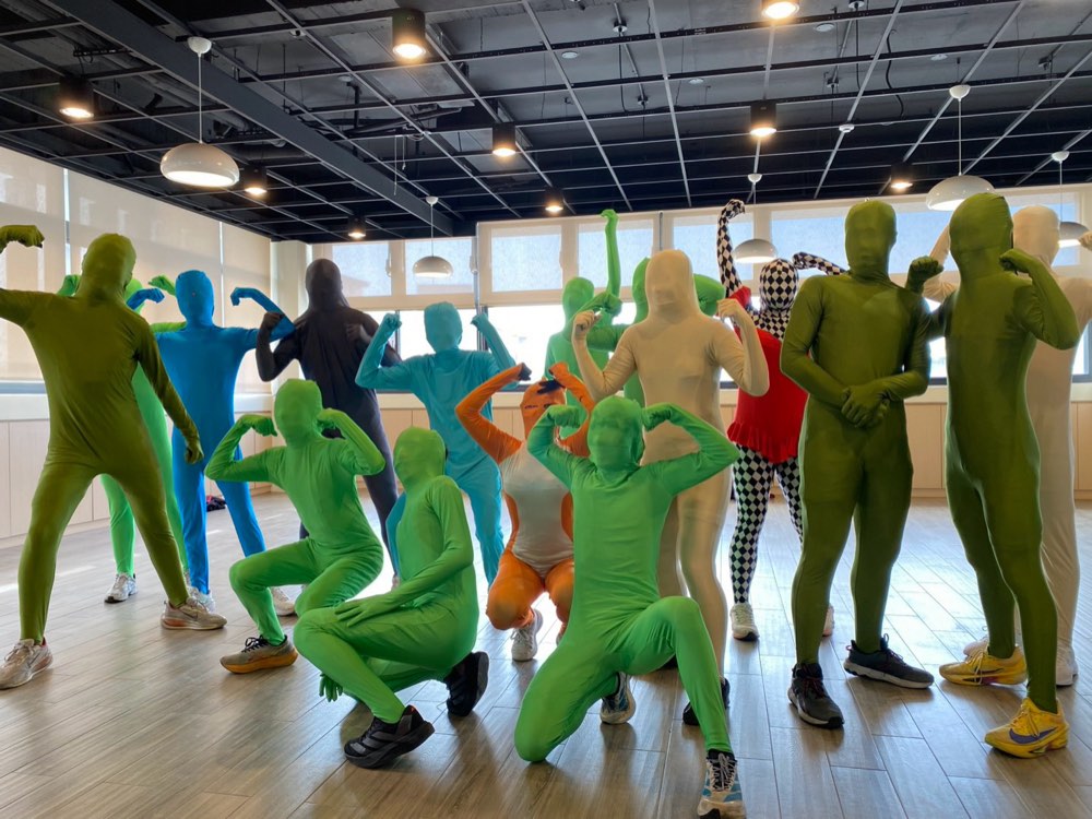 Students in green, blue, orange and checkered bodysuits strike poses together indoors