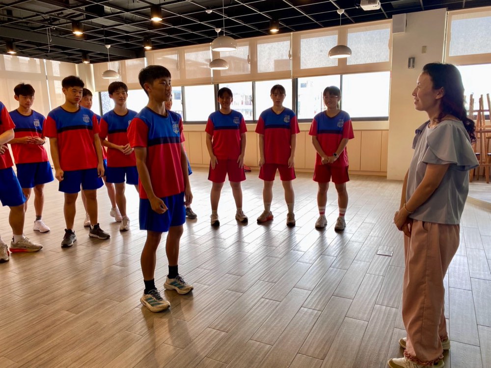 Students in sports uniforms stand in a circle in the studio, listening for the next instruction