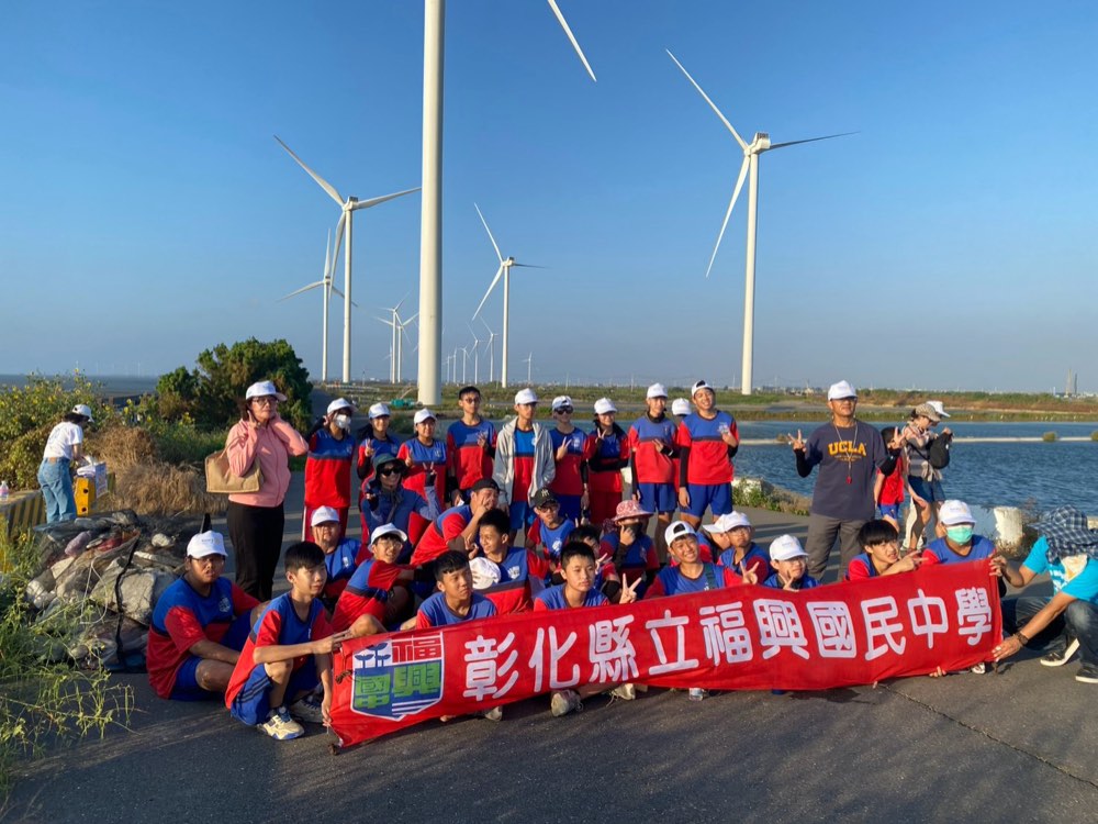 Fusing students and teachers holding the school banner beneath wind turbines at Hanbao Wetland
