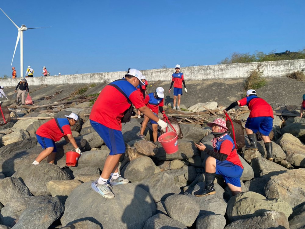 Students in gloves and rain boots picking up trash among the seawall rocks, buckets in hand