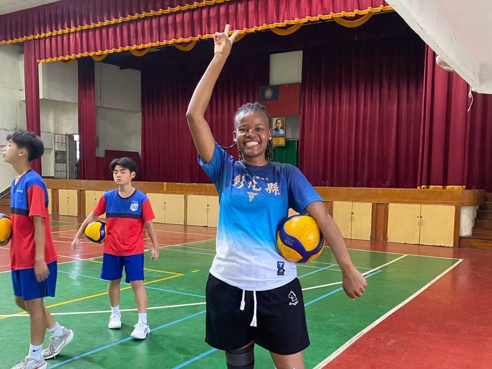 Teacher Z smiles and flashes a peace sign with a volleyball under her arm in the school gym
