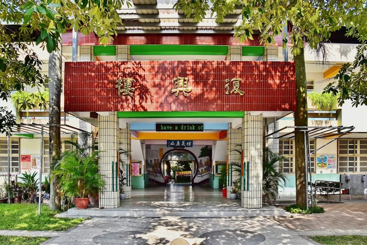 復興國小的復興樓：兩根白色磁磚方柱撐起門廊，上方是暗紅色磁磚牆面，嵌著由右至左的金色大字「復興樓」，門楣下掛著一台跑馬燈字幕機，正顯示英文字句，門內看得見「禮義廉恥」匾與圓形的勤學門，兩側是棕櫚與大樹，樹影落在紅牆上 The 復興樓 building at Fuxing — two white-tiled piers carrying a porch, a dark red tiled panel above set with gold characters reading 復興樓, an LED ticker under the lintel showing a line of English, the 禮義廉恥 plaque and the round 勤學門 visible through the opening, palms and large trees on both sides casting shadows across the red wall