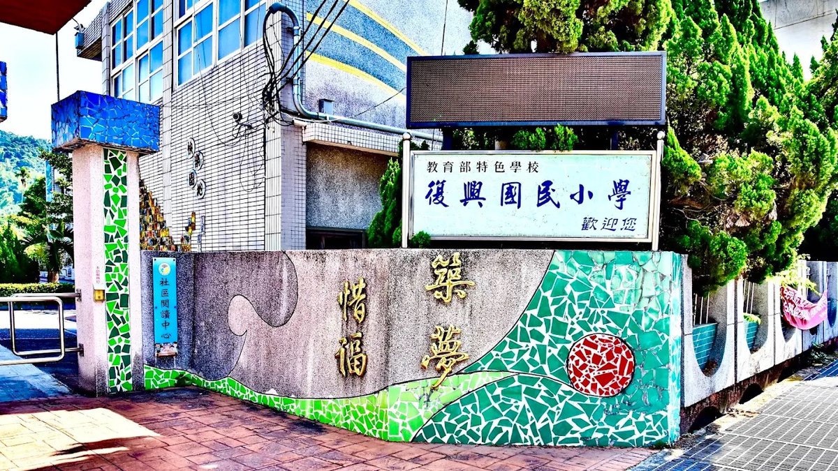 復興國小校門旁的照壁：洗石子牆面嵌著金色大字「惜福築夢」，右半邊是藍綠色碎磁磚拼成的水波與一輪紅色的日，牆上方掛著白底藍字的校名牌，上排小字寫著「教育部特色學校」，下排寫著「歡迎您」，牆邊立著一塊「社區閱讀中心」標示牌 The wall beside the Fuxing gate — gold characters reading 惜福築夢 set into washed stone, the right half tiled in green and blue mosaic as water with a red mosaic sun in it, a white name board above reading 教育部特色學校 復興國民小學 歡迎您, and a small sign at the left reading 社區閱讀中心