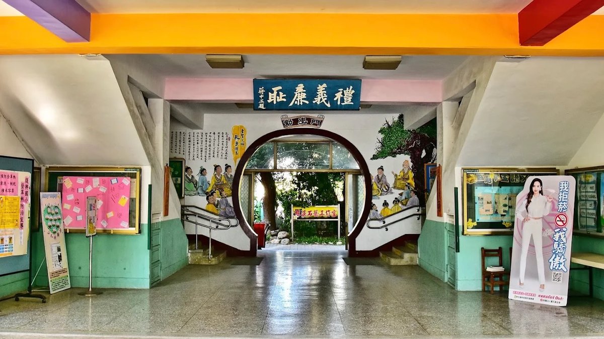復興國小穿堂：磨石子地面反射著光，正中央是勤學門的月洞門，門上方牆面掛著藍底白字的匾額「禮義廉恥」，天花板橫樑漆成黃、紫、粉、紅，兩側牆下段是墨綠色牆裙，左右各有玻璃公佈欄與海報，右側立著一塊宣導立牌 The entrance hall at Fuxing — polished terrazzo reflecting the light, the round 勤學門 opening in the middle, a blue plaque in white characters reading 禮義廉恥 on the wall above it, ceiling beams painted yellow, purple, pink and red, dark green dado along the side walls, glazed noticeboards and posters on both sides and a free-standing campaign board at the right