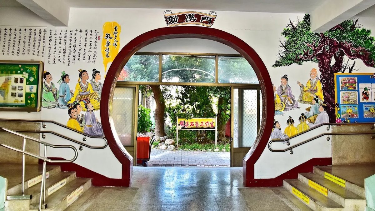 復興國小的勤學門：白牆上開著一道圓形的月洞門，門框漆成深紅色，上方掛著「勤學門」匾額，左右牆面彩繪孔子講學的場景&mdash;&mdash;弟子們穿黃、綠、紫衣裳環坐聆聽，右側畫著一棵老松與孔子，左側牆上有一段介紹孔子的文字與一塊標題為 English Column 的公佈欄，門內看得出去是綠樹與一塊木招牌 The 勤學門 moon gate at Fuxing — a circular opening in a white wall framed in deep red, a plaque reading 勤學門 above it, murals of Confucius teaching on either side with students in yellow, green and purple robes seated around him, a pine tree painted at the right, a panel of text about Confucius and a glazed noticeboard headed English Column at the left, and green trees and a timber sign visible through the opening
