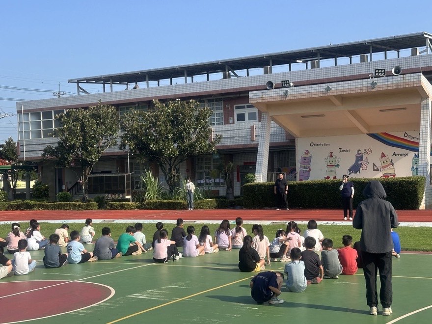 朝會全景，學生坐在球場地面上，師長在司令台前拿著麥克風說話，背景是彩虹牆與藍天
