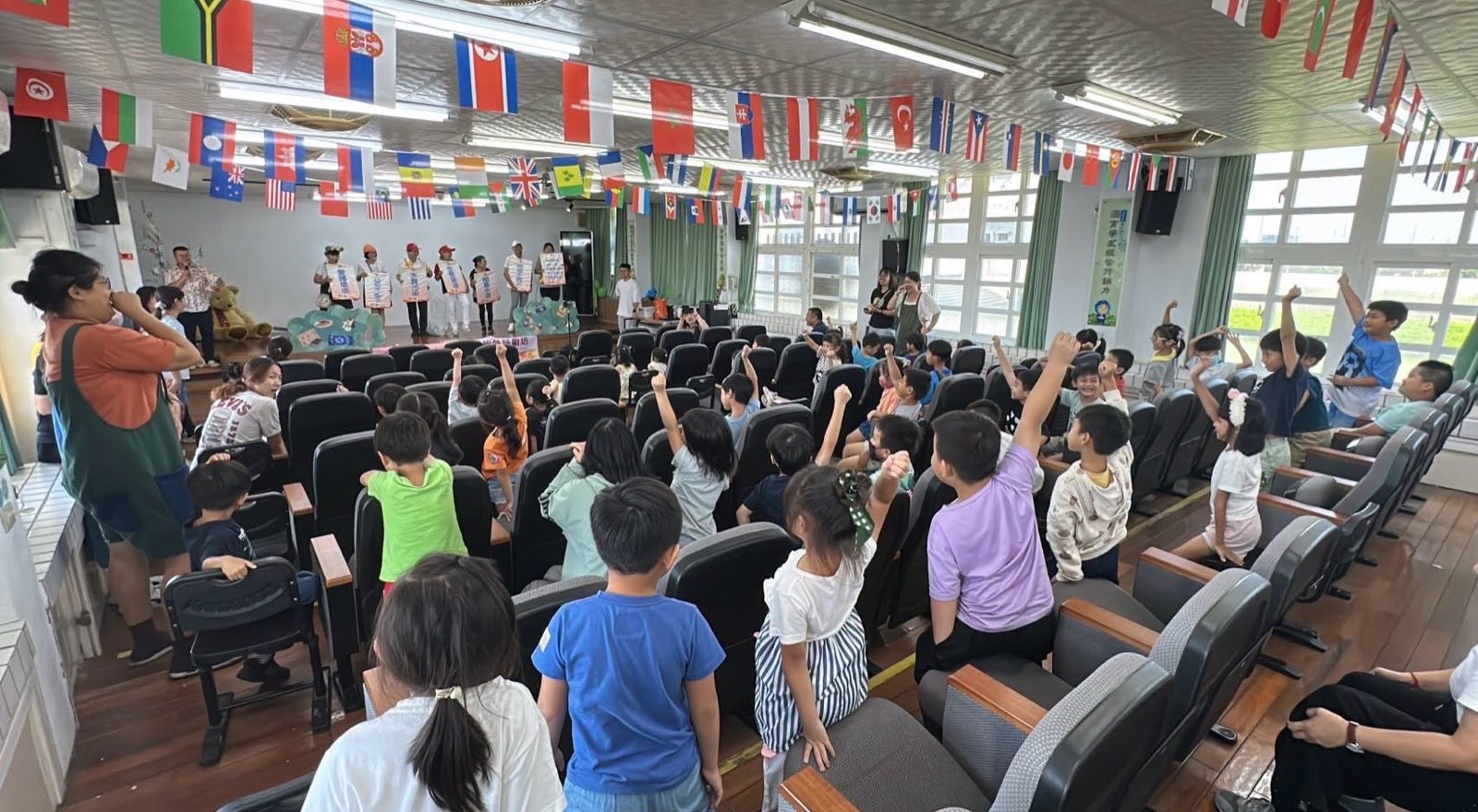 視聽教室裡，台上演員舉著環保標語牌，台下學生高高舉手搶答，天花板掛滿萬國旗