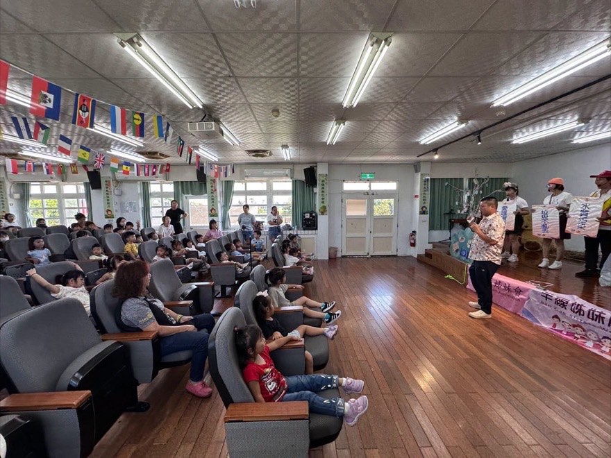 台上演員分別舉著「愛護環境」「垃圾不亂丟」「水質污染」標語牌，幼兒園小朋友坐在觀眾席觀賞
