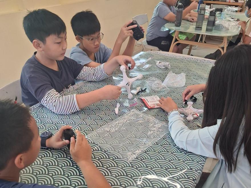 學生圍著桌子分組組裝，手上拿著白色無人機機身和小零件