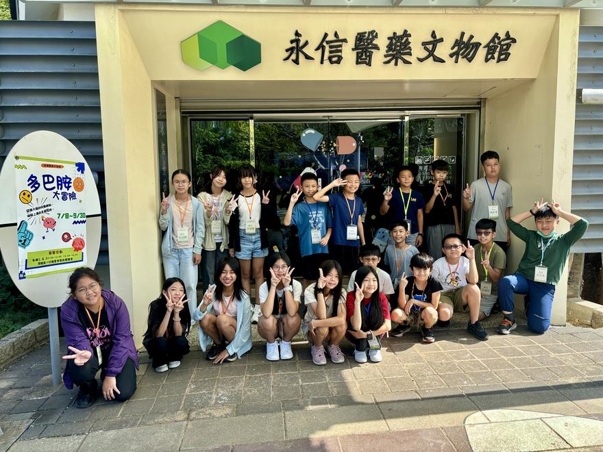 六年甲班學生在永信醫藥文物館入口前比 YA 合照，左側立牌是「多巴胺大冒險」特展