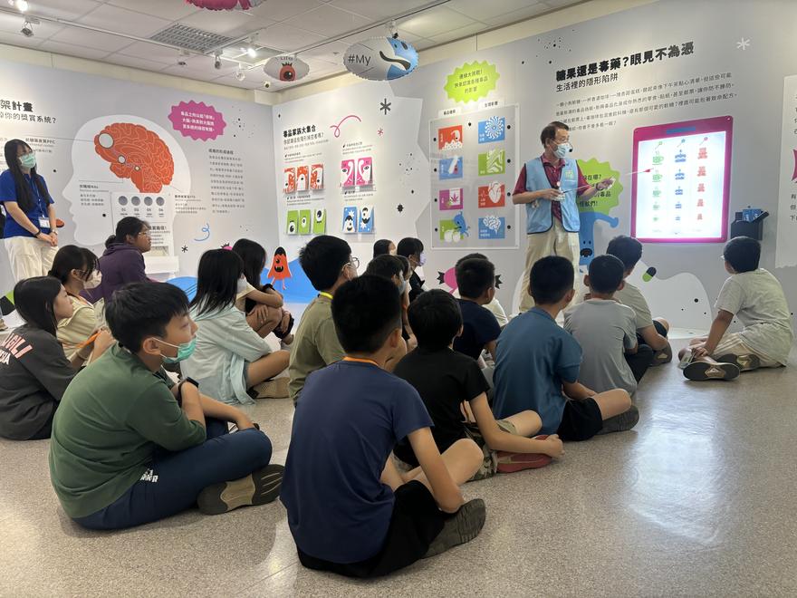 學生席地而坐，志工導覽員在「糖果還是毒藥？眼見不為憑」展板前解說