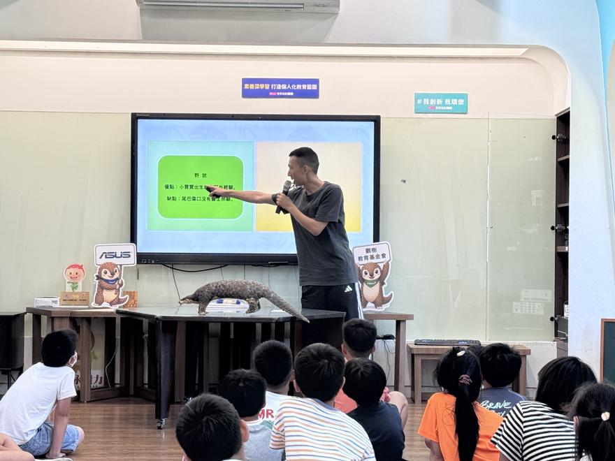 講師指著螢幕上的簡報說明，桌上擺著一隻穿山甲模型