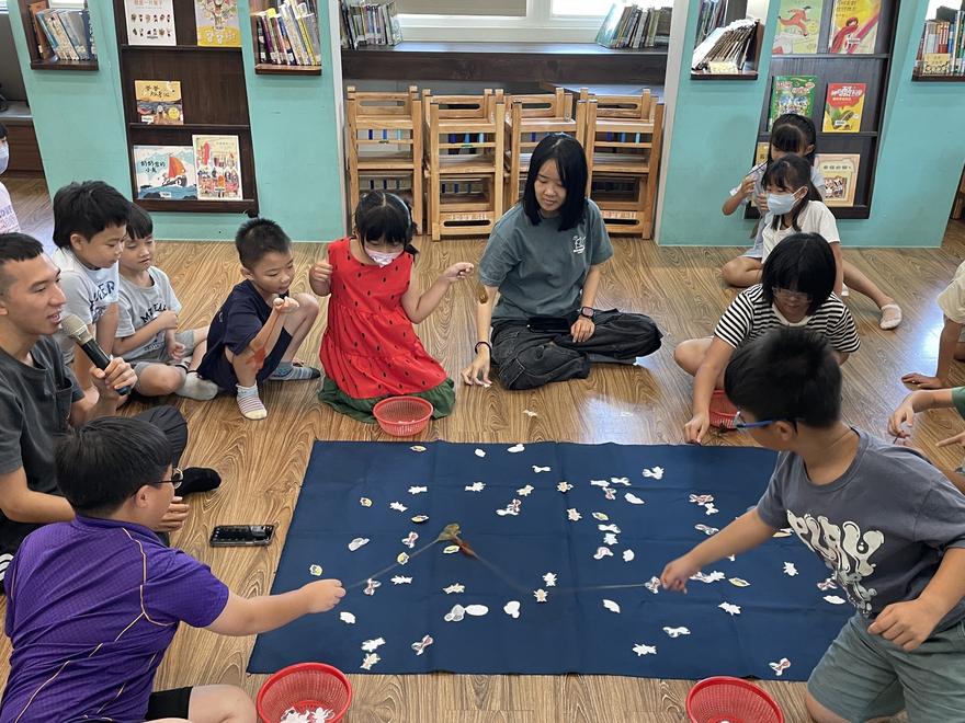 學生圍坐在藍色地墊旁，拿著長棒子挑起地墊上的紙片小蟲