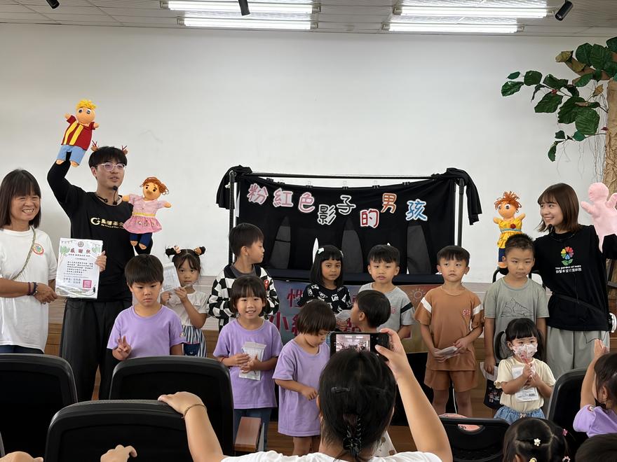 演員舉起布偶，與穿紫色上衣的幼兒園小朋友在「粉紅色影子的男孩」偶台前合影
