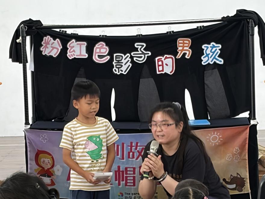 偶台前，一位小男孩拿著紙張上台分享想法，主持人在一旁拿麥克風協助