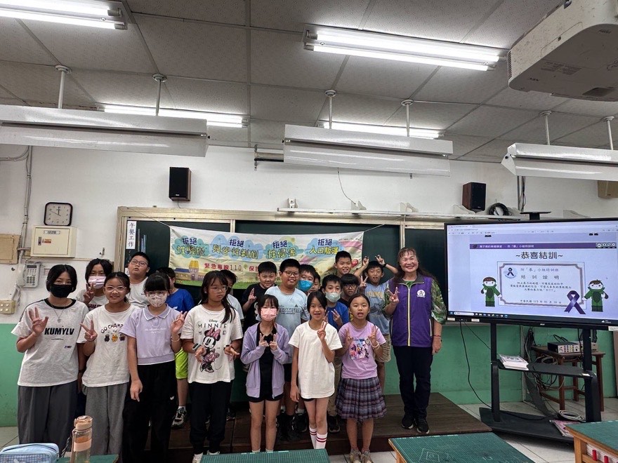 高年級學生與講師在教室合影，螢幕上顯示防暴小組特訓班的結訓畫面