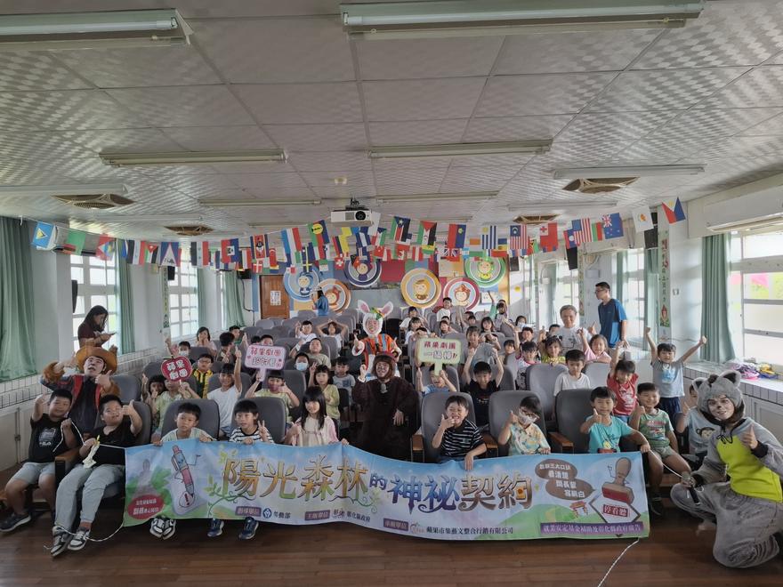 全校師生與動物角色在禮堂大合照，前排拉著「陽光森林的神祕契約」布條，布條右側寫著勞動三大口訣