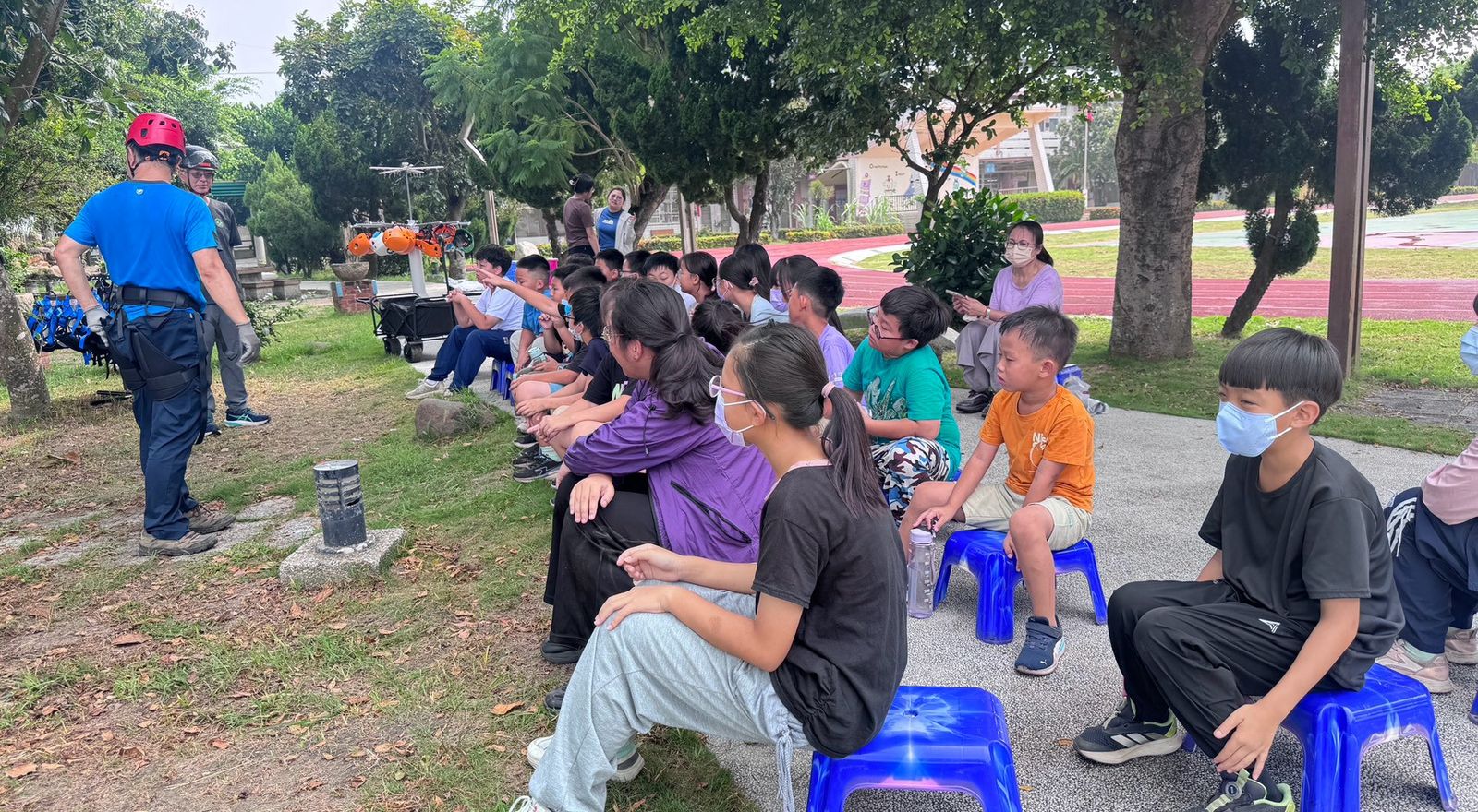 廣興國小學生坐在校園大樹下，聽攀樹教練講解安全裝備與上升技巧