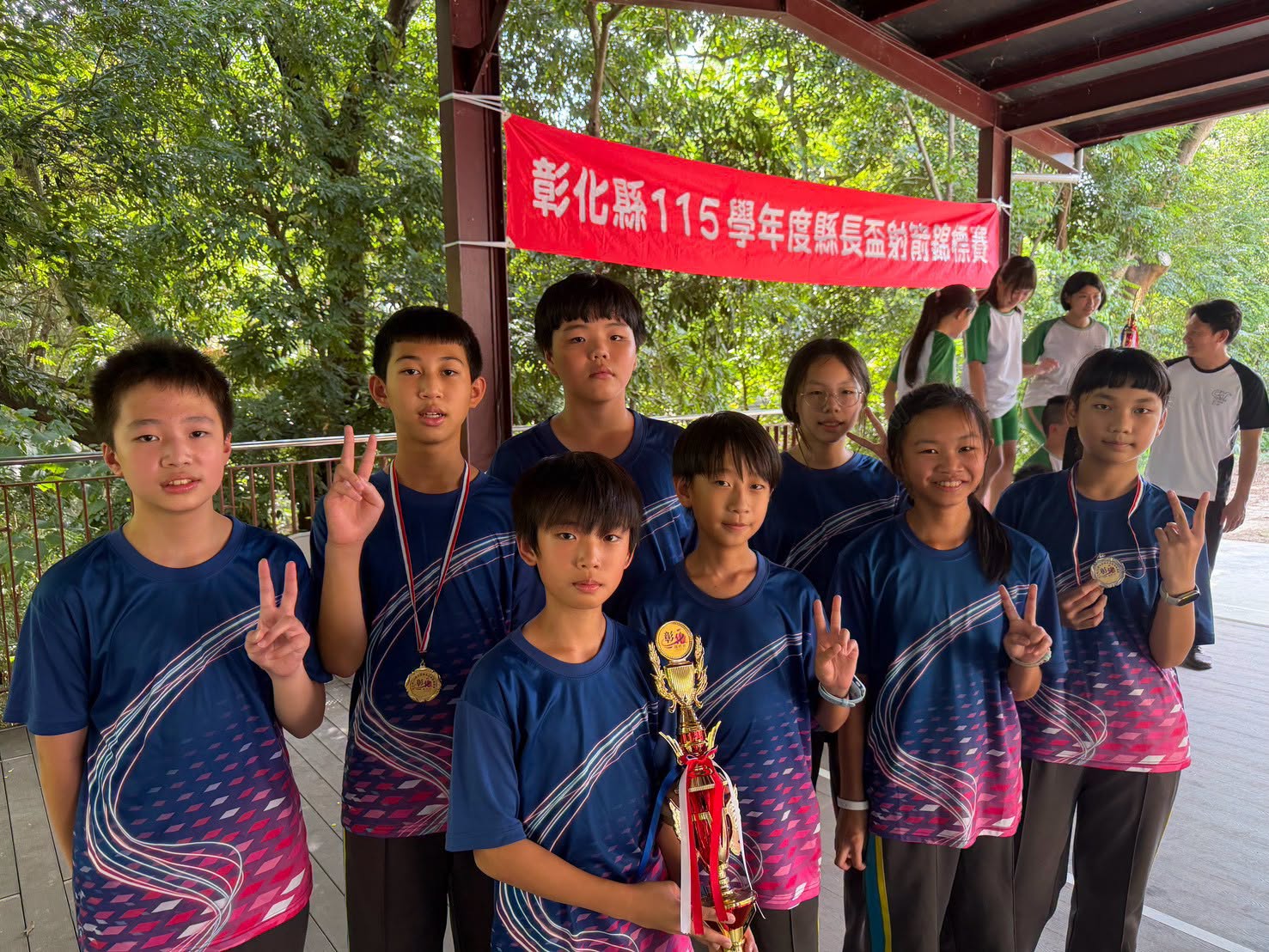 新民國小射箭隊選手戴著獎牌、捧著亞軍獎盃，在縣長盃射箭錦標賽布條前合影