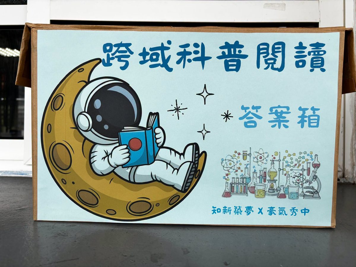 「跨域科普閱讀 答案箱」紙箱，上面畫著坐在月亮上看書的太空人，下方寫著「知新築夢 X 豪氣秀中」 The Cross-Disciplinary Science Reading answer box, decorated with an astronaut reading a book on the moon