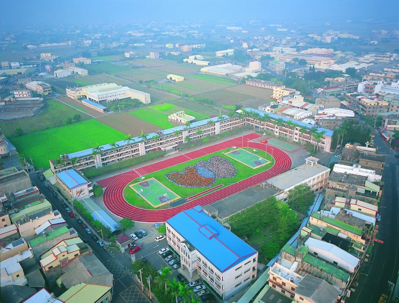 從空中俯瞰秀水國中：紅色跑道圍著綠色操場，四周是校舍與秀水鄉的田野和房舍 An aerial view of Hsiushui Junior High: a red running track around a green field, with school buildings, farmland and the houses of Xiushui all around