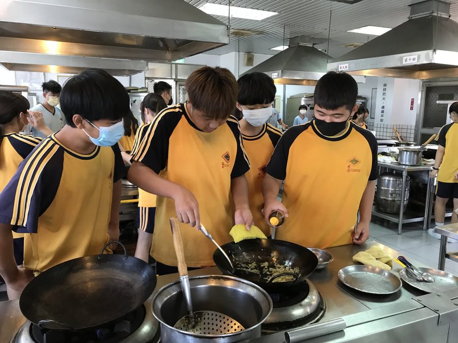 秀水國中學生在專業廚房裡圍著爐台，用炒鍋練習烹調 Hsiushui students around a stove in a professional kitchen, practicing cooking with woks