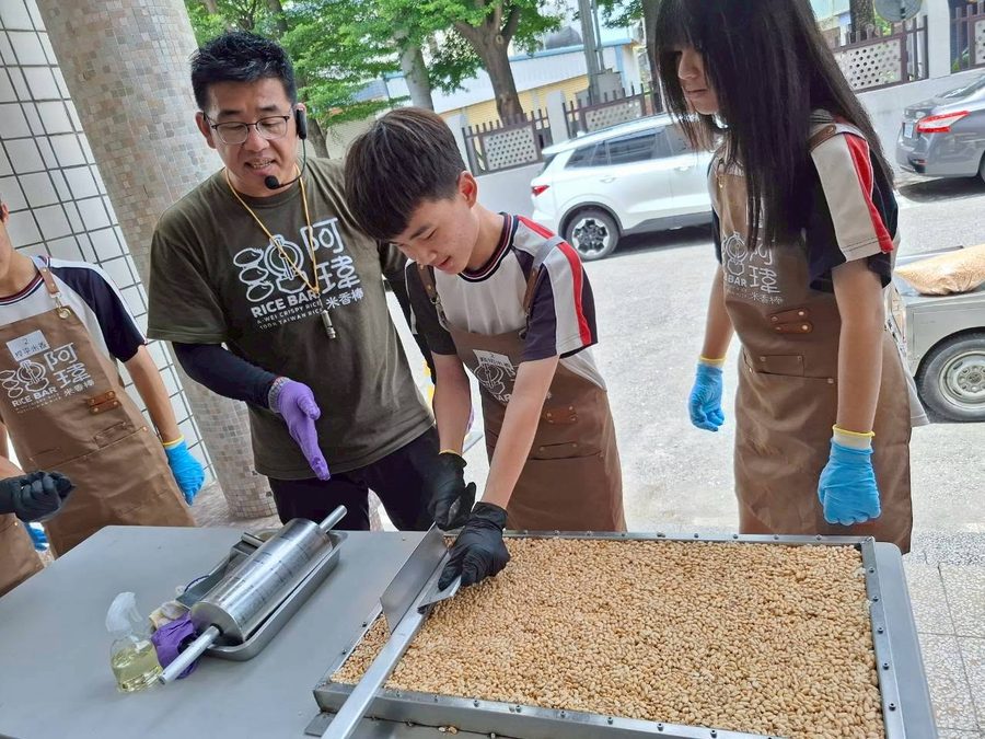 米香職人示範切米香，秀水國中學生戴著手套在一旁學習 A puffed-rice bar maker shows Hsiushui students how to cut a tray of rice bars