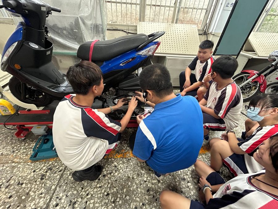秀水國中學生蹲在機車旁，跟著老師學習機車修護 Hsiushui students crouching beside a scooter, learning scooter maintenance from a teacher
