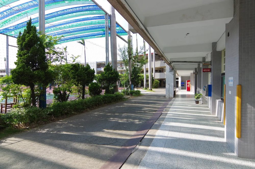 秀水國中校舍旁的長廊，左側是藍綠色遮陽棚與樹木 A long walkway beside a Hsiushui school building, with blue-green shade canopies and trees on the left