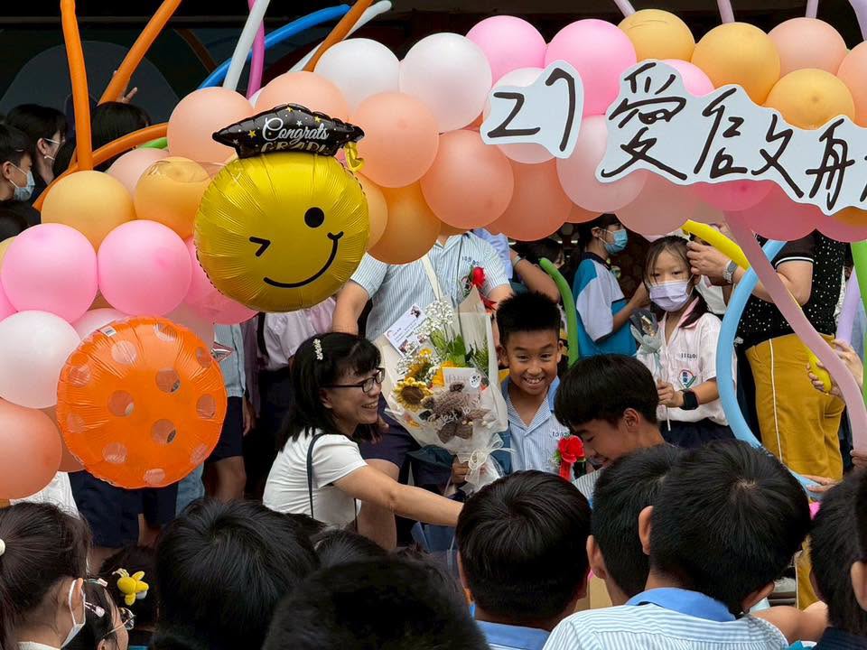 A graduate receiving flowers under a balloon arch reading '27'