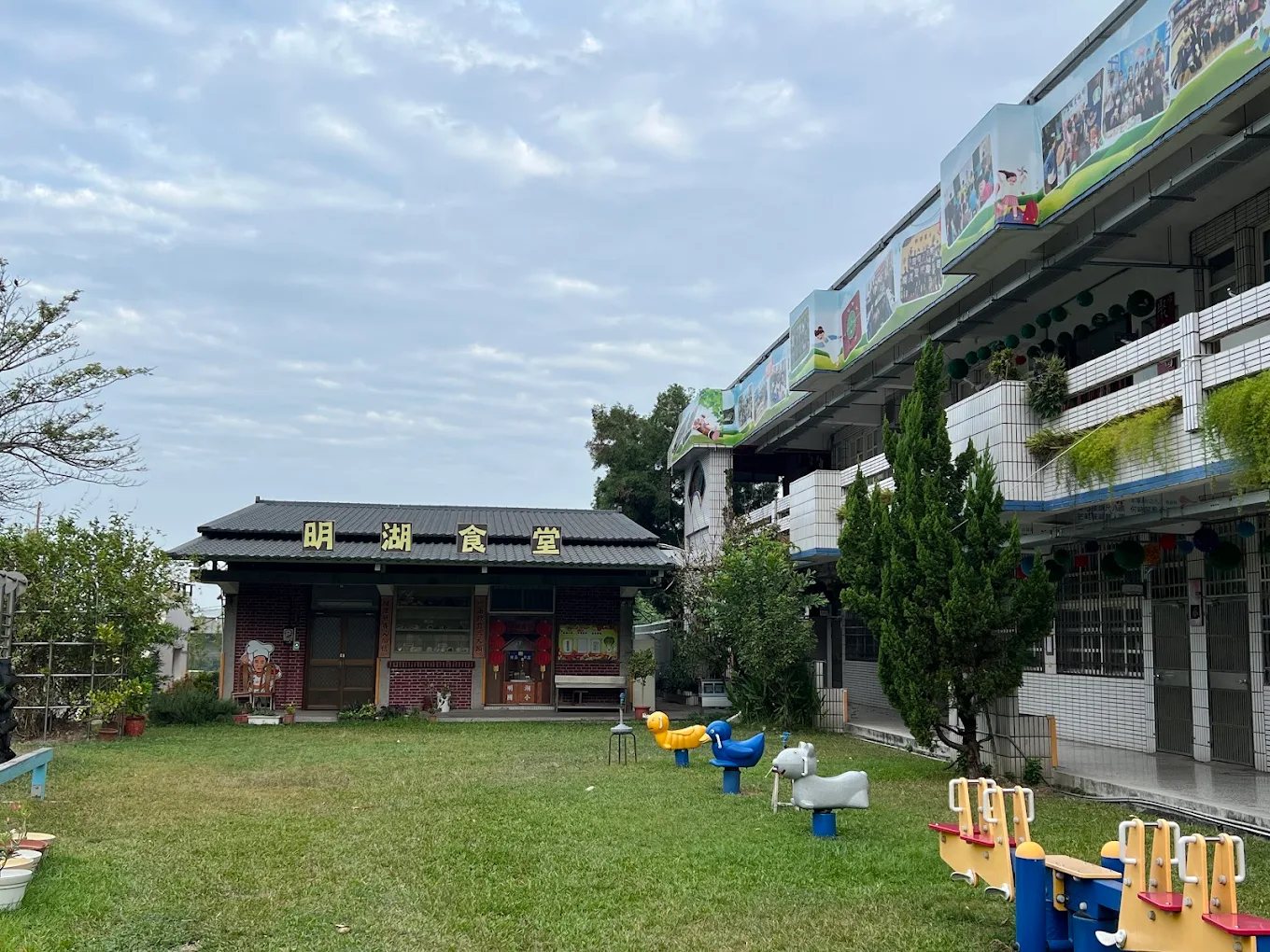 明湖國小的草地與「明湖食堂」：黑瓦紅磚的平房上以金字寫著食堂名稱，草地上散著鴨子、牛與小馬造型的彈簧搖搖馬與蹺蹺板，右側是三層樓教室，欄杆上掛著一排學生活動照片看板 The lawn at Minghu with the 明湖食堂 dining hall — a low red-brick building with a black tiled roof and the hall's name in gold characters, spring rockers shaped like ducks, a cow and a pony scattered across the grass, and the three-storey classroom block on the right with a row of photo boards of student activities along its railing