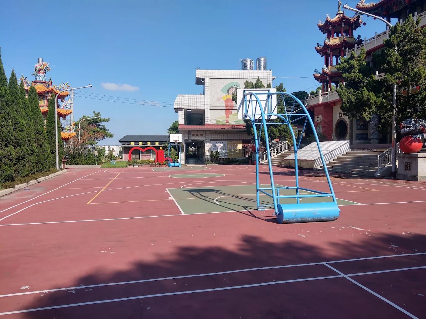 明湖國小的紅色球場，兩側各立著開林寺的彩繪樓閣與龍柱，中央是藍色籃球架與司令台，遠處可見紅色心形拱門，藍天無雲 The red-surfaced ball court at Minghu, flanked on both sides by the painted towers and dragon columns of Kailin Temple, a blue basketball frame in the middle and the red heart arch visible in the distance under a clear blue sky