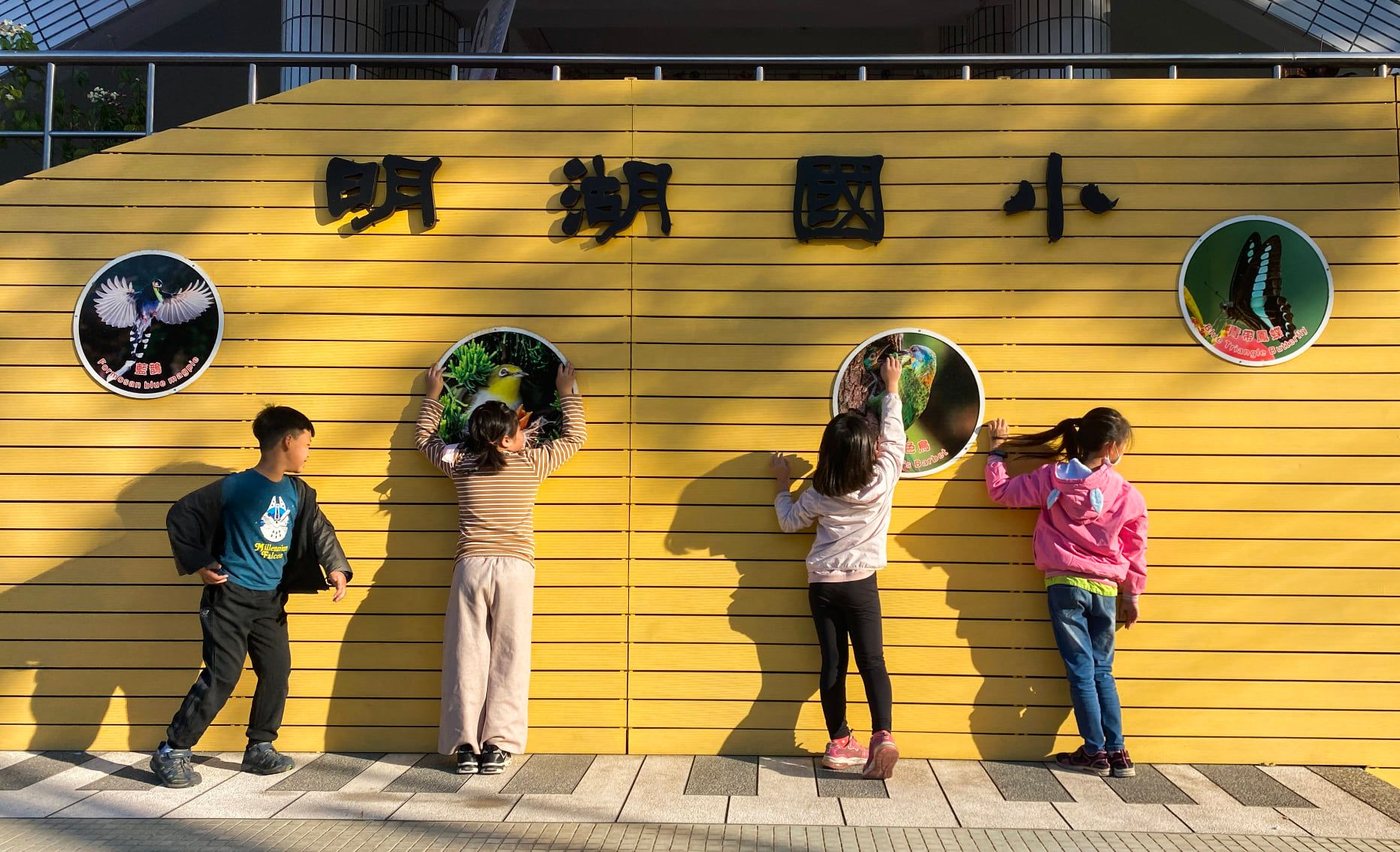 四個孩子背對鏡頭，站在明湖國小黃色的圍牆前，伸手摸牆上的圓形生態解說牌；牆上有「明湖國小」四個黑色大字，圓牌上印著台灣藍鵲、五色鳥、綠繡眼與青帶鳳蝶的照片與中英文名稱 Four children with their backs to the camera, standing at the yellow wall of Minghu Elementary and reaching up to touch the round ecology plaques mounted on it; the school name is spelled out in large black characters above, and each plaque carries a photograph of a Formosan blue magpie, a Taiwan barbet, a white-eye or a blue triangle butterfly with its name in Chinese and English