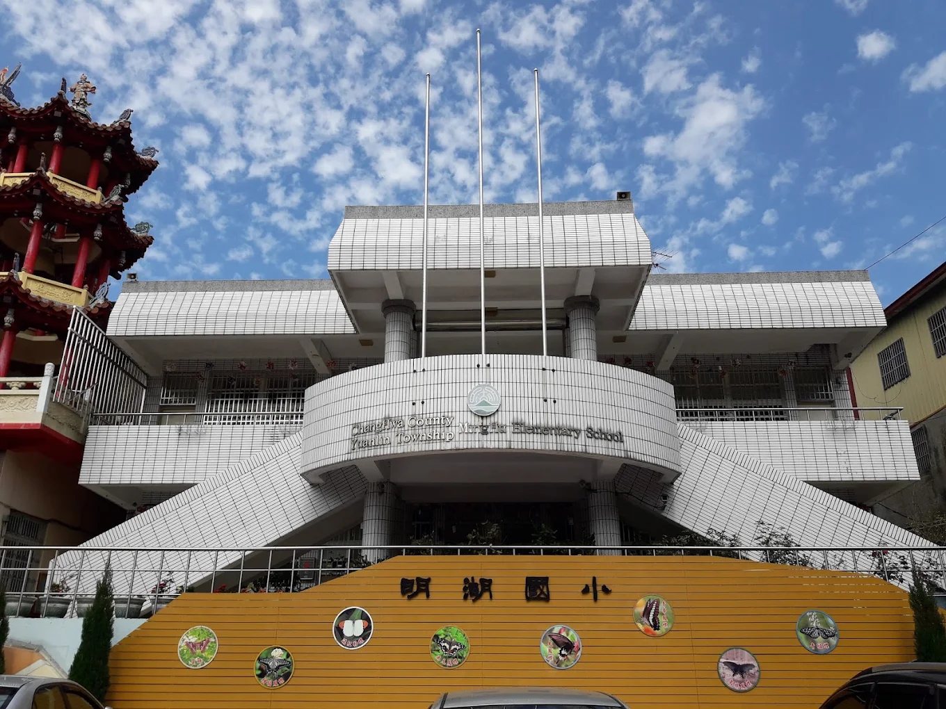明湖國小主校舍正面：白色三層樓校舍與弧形門廊，左側緊鄰開林寺紅柱金瓦的樓閣，前方黃色圍牆上有「明湖國小」四個大字與一排生態圓牌，藍天白雲 The front of the main classroom block at Minghu — a white three-storey building with a curved portico, the red pillars and golden tiles of Kailin Temple immediately to its left, and the yellow wall in front carrying the school name and a row of round ecology plaques under a cloudy blue sky