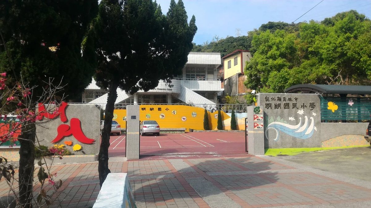 從校門外望向明湖國小：白色校舍後方是一整片林木蒼翠的山坡，左側有一株盛開的山櫻花，門柱上是紅色的校徽字樣與金色校名 Looking in from outside the Minghu gate — the white classroom block with a densely wooded hillside rising behind it, a mountain cherry in bloom on the left, and the red crest lettering and gold school name on the gate piers